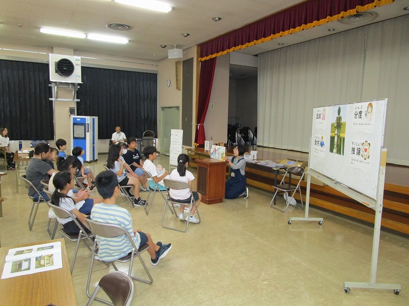 子供たちと数名の保護者がパイプ椅子に座って、ホワイトボードの文字やイラストを使いながら説明する女性講師の話を熱心に聞いている写真