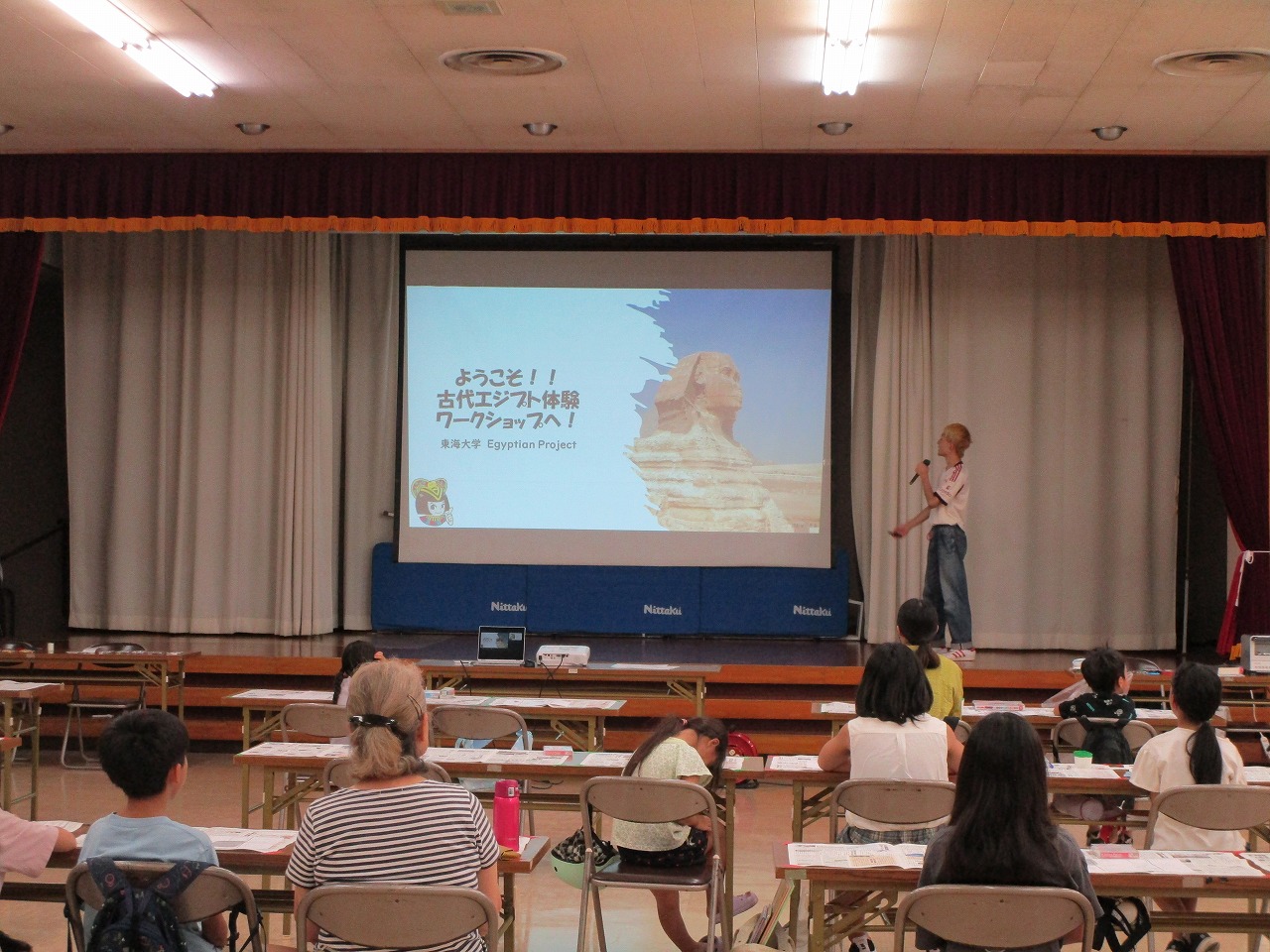 大きなスクリーンに「ようこそ!! 古代エジプト体験ワークショップへ! 東海大学 Egyptian Project」の文字が映し出される中、男性がマイクを持ち聴衆に向かって話している講演の様子の写真