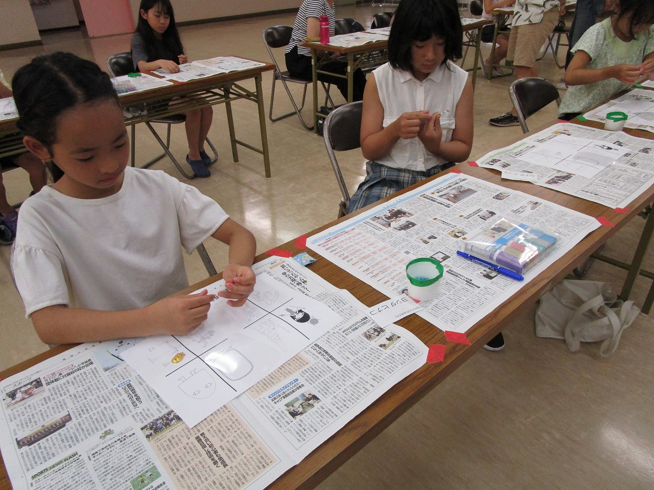 広間で新聞紙と白い紙で覆われた長机に座った複数の子供たちのうち、手前の黒い三つ編みの女の子が白い紙の上で何かを作成し、その奥の女の子も手元に集中しているワークショップの様子の写真