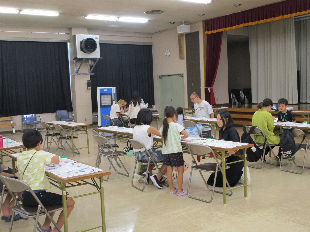 子供たちが長机と折りたたみ椅子に座り、数人の大人と一緒に学習や作業をしている様子を捉えた屋内写真