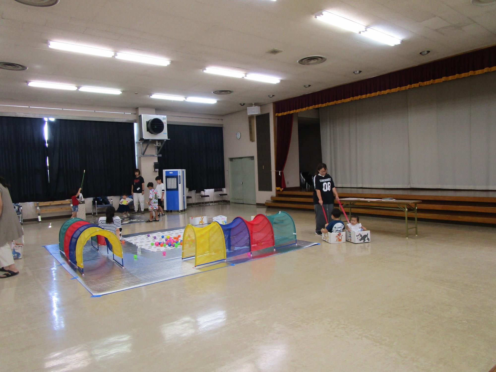 ビニールシートの上にボールプールやトンネルのおもちゃが置かれ、複数の子供たちが遊び、一人の大人が段ボールに乗せた子供たちを押して遊んでいるレクリエーションの様子の写真