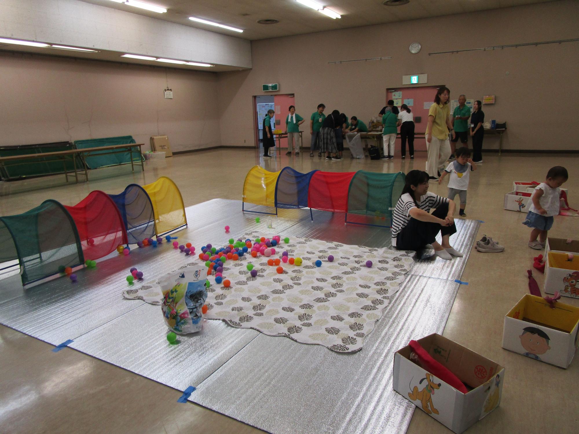 銀色のシートの上に色とりどりのボールが散らばるボールプールとトンネルの遊具が設置され、数名の親子と緑のポロシャツを着たスタッフが立っているレクリエーションの様子の写真