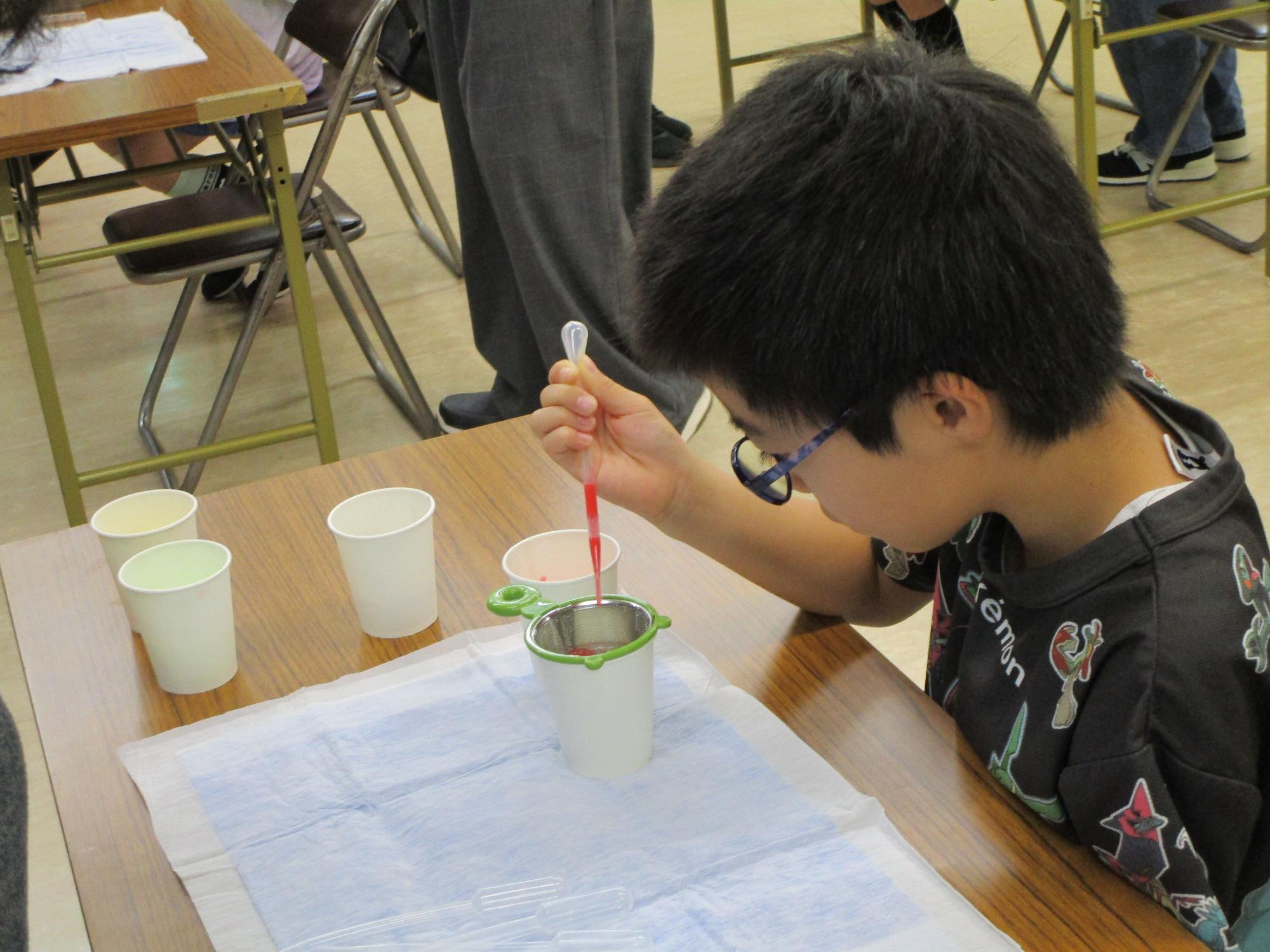 眼鏡をかけた男の子が、机の上の白い紙コップと青い吸収シートの上で、スポイトを使い赤い液体を小さなザルに入れたコップに滴下している実験の様子の写真