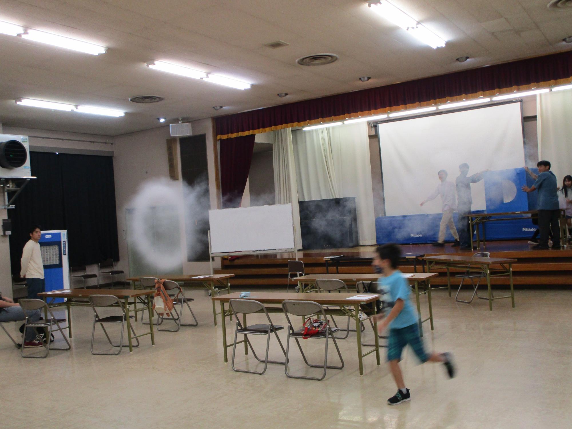 ドライアイスを使った煙の実験で出た空気砲による煙の輪を青いシャツを着た男の子が追いかけている様子の写真