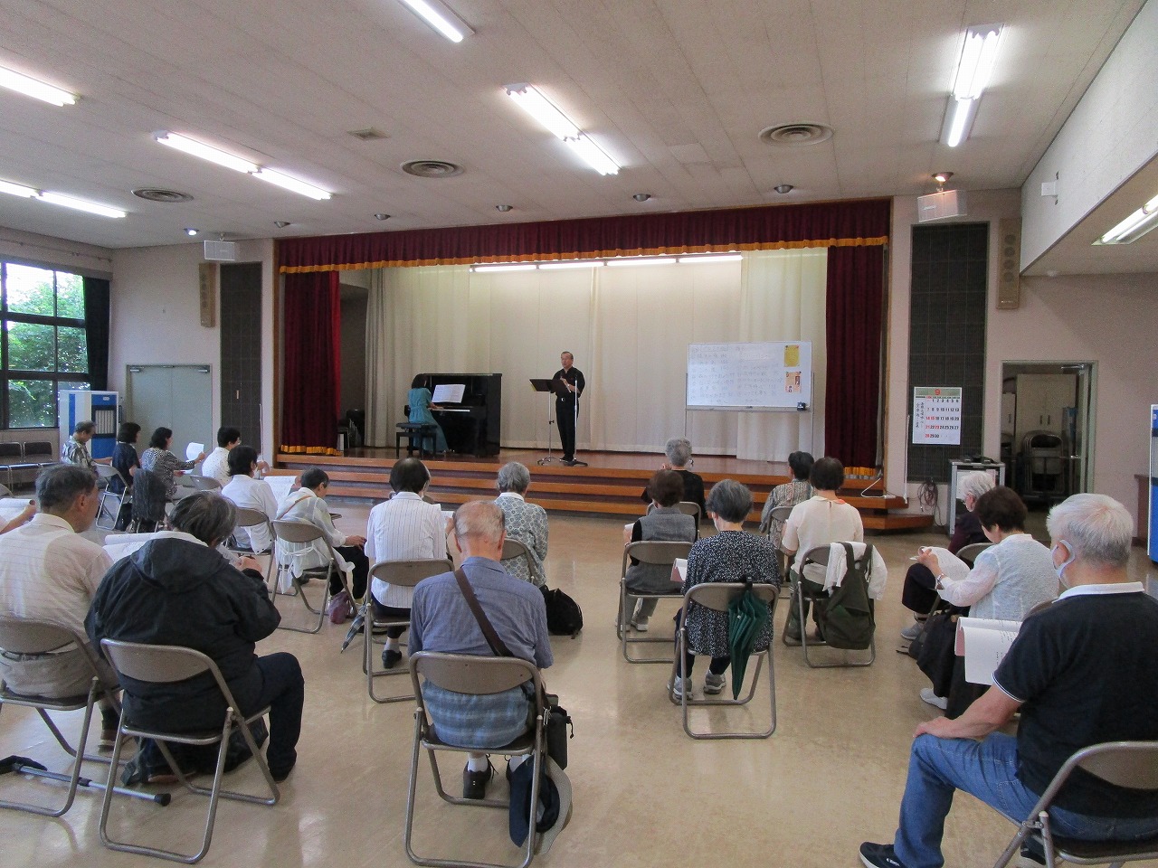 ホールのような広い会場で、折りたたみ椅子に座った高齢者を中心とした聴衆がステージ方向を向いており、ステージ上では女性がピアノを演奏し、男性がマイクを使って聴衆に向かって立って話している様子の写真