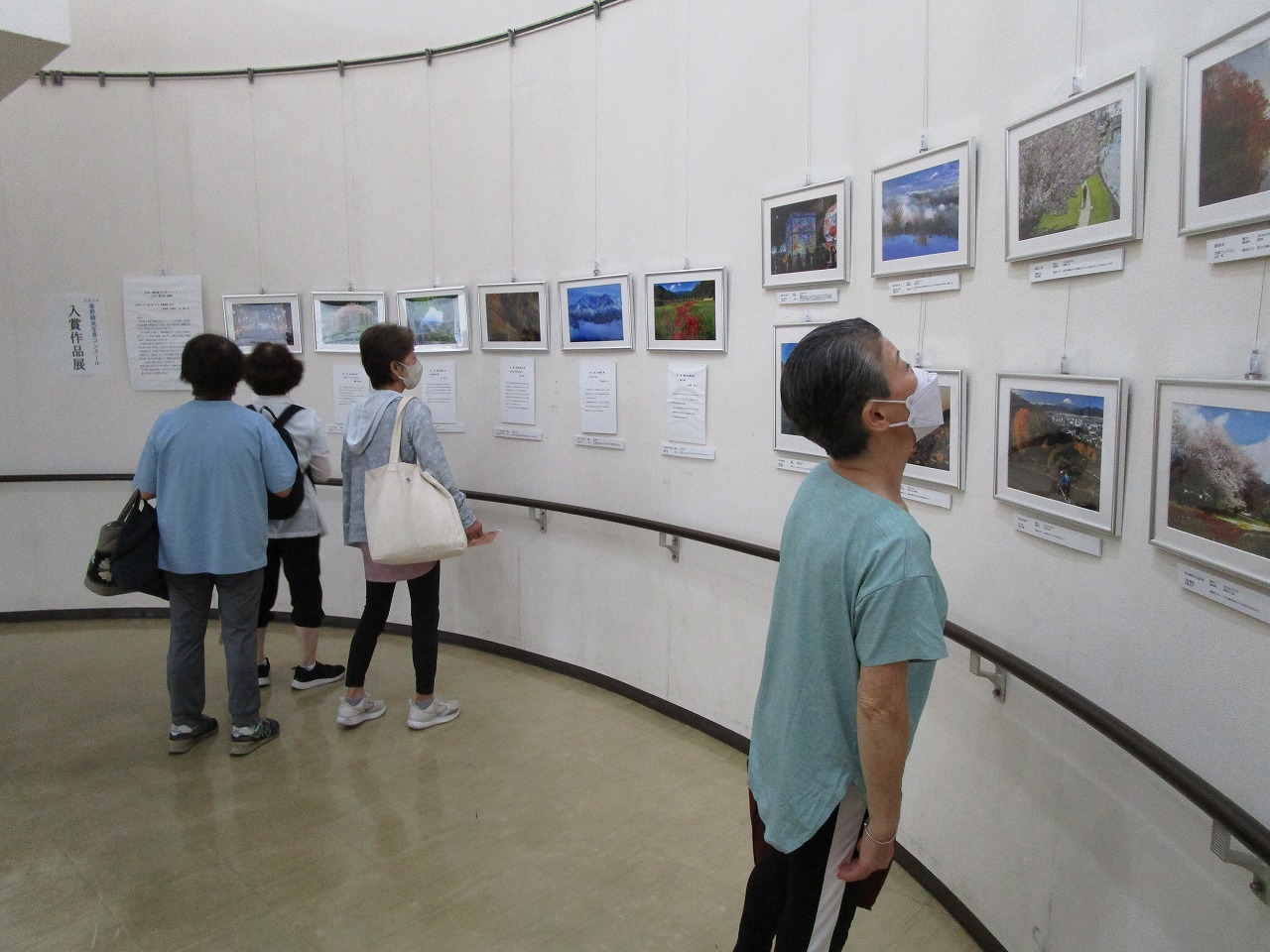 曲面を描いた白い壁に風景写真が多数展示された会場で、マスクを着用した4人の女性たちが熱心に作品を鑑賞している写真