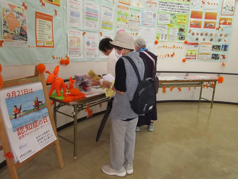 世界アルツハイマーデーに関連したポスターや情報掲示がされた壁の前で、数人の高齢の女性たちがテーブルに置かれた資料を読んだり、熱心に見ている様子の写真