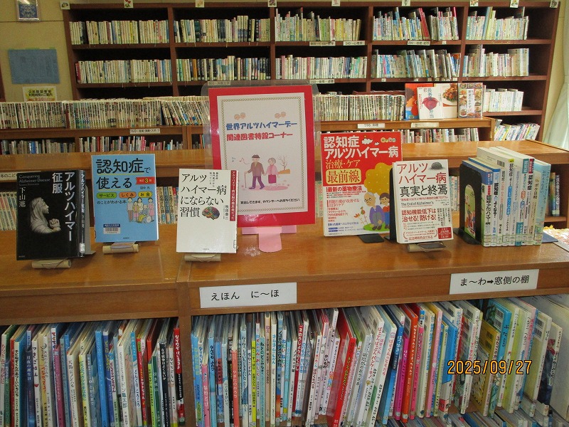 図書室の本棚の上に、「世界アルツハイマーデー関連図書特設コーナー」のポスターが中央に立てられ、その両脇に認知症やアルツハイマー病に関する複数の啓発本や専門書が並べられている写真