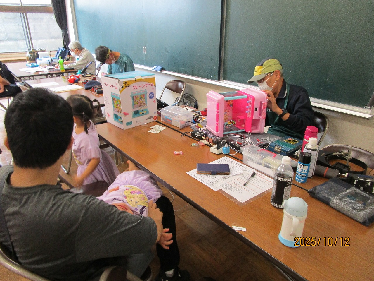 男性が机に向かい、おもちゃを修理し、手前では父親が子供を膝に乗せて待っている写真