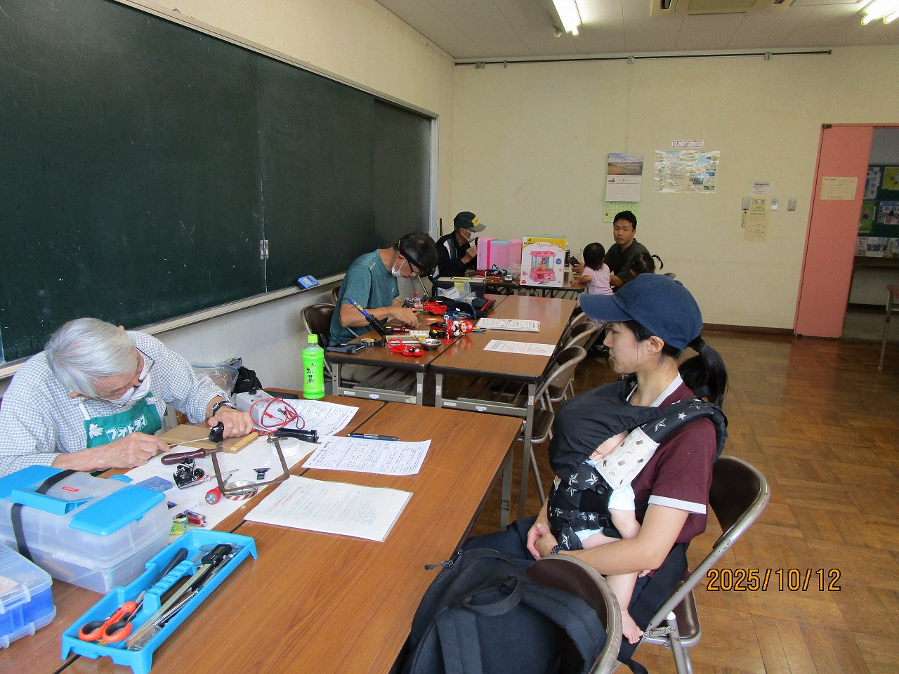 手前の高齢男性が道具を使って作業に集中し、赤ちゃんを抱っこして座っている女性が作業を見ている写真