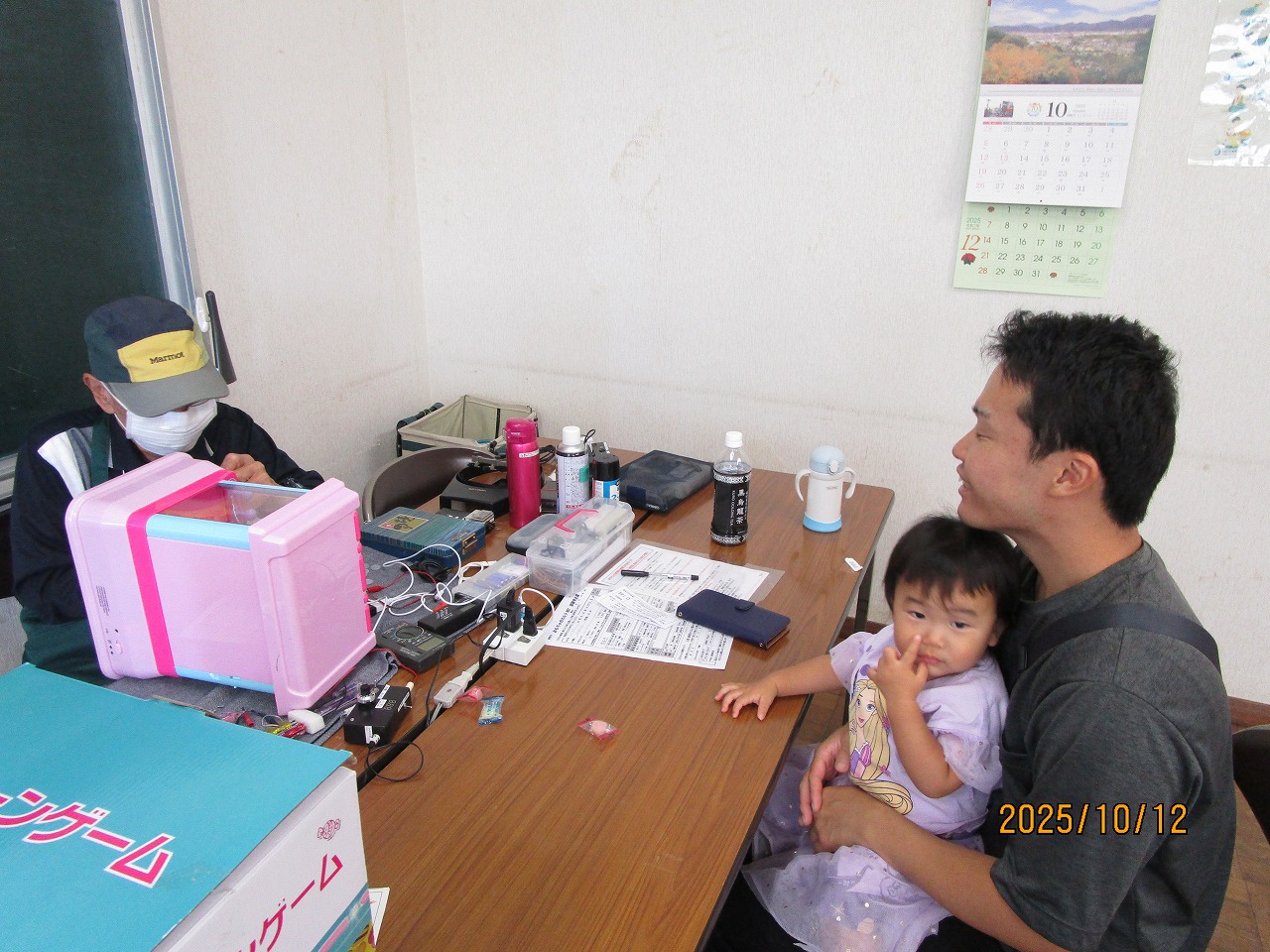 机で作業する男性と、手前の机で幼い子供を膝に乗せて見守る男性の親子が、おもちゃを修理しているのを見ている写真