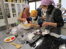 調理室のテーブルの上で、二人でケーキの材料をミキサーで混ぜている、一人はそれを見ている。