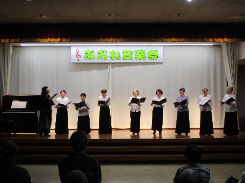 ステージ上で女性数名が白のブラウス黒のスカートで譜面を持ちながらコーラスを披露している