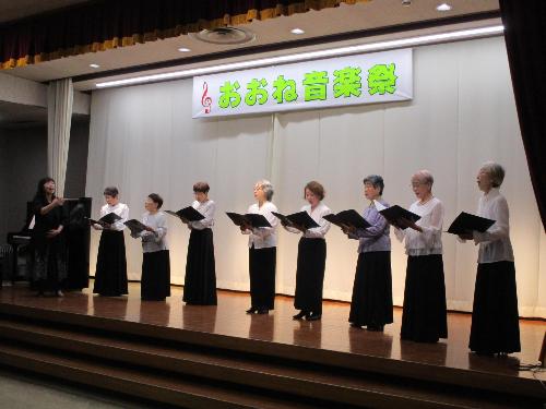 ステージ上で女性数名が白のブラウス黒のスカートで譜面を持ちながらコーラスを披露している