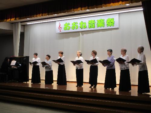 ステージ上で女性数名が白のブラウス黒のスカートで譜面を持ちながらコーラスを披露している。一人はピアノで伴奏している。