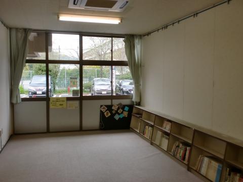 正面のガラス窓から外光が差し込んでいて、右側の白い壁際に2段の書棚があり半分ほど本が入っていて、床はカーペット敷きの児童室の写真