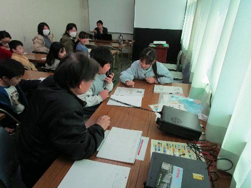 男の子が無線機のマイクに向かって話しかけているのを、十数人の受講者が見ている様子