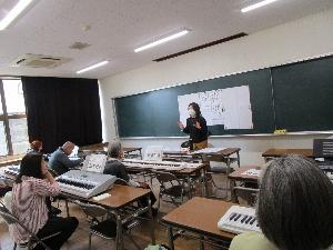 黒板に資料が貼られていて、講師が受講者に説明を行っている。受講者数人はそれぞれキーボードを置いた机に向かって座っている。