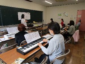 数人の受講者がそれぞれの机の上のキーボードを操作していて、講師が黒板に貼った資料で説明をしている。