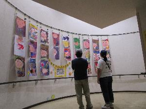 水彩画で顔を書いた画用紙二十数枚が壁面に展示されているのを二人の大人が鑑賞している様子