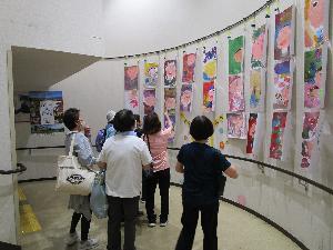 水彩画で顔を書いた画用紙二十数枚が壁面に展示されているのを大人数人が鑑賞している様子