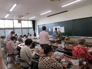 教室で十数人の生徒がテーブルの上で針仕事をしている様子、一人の生徒が講師に尋ねている。