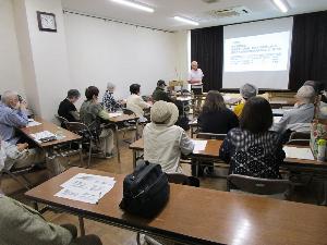 講師が資料をプロジェクターに映し説明しているのを10数名の受講者が聴いている。