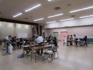 複数のテーブルそれぞれで、数人の小学生が粘土で工作をしているのを数人の大人が指導している様子