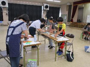 複数のテーブルそれぞれで、数人の小学生が粘土で工作をしているのを数人の大人が指導している様子