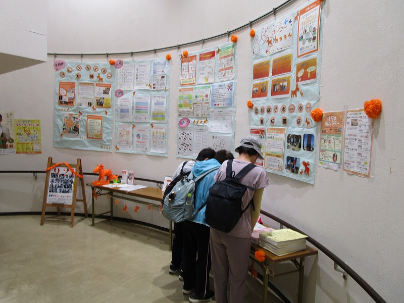 壁に資料が展示されていて、テーブル上に資料が置かれていて、大人数人がテーブル上の資料を見ている