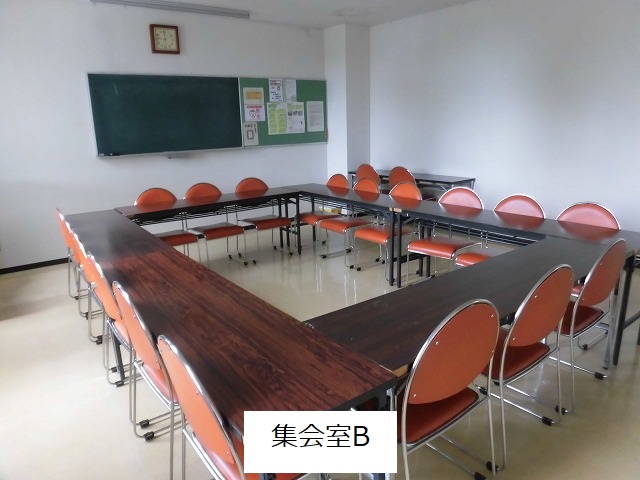 白い壁の室内で、ダークブラウンの長机とオレンジ色の背もたれを持つ椅子がロの字型に並べられ、奥の壁には黒板と掲示板が備え付けられている集会室Bの写真