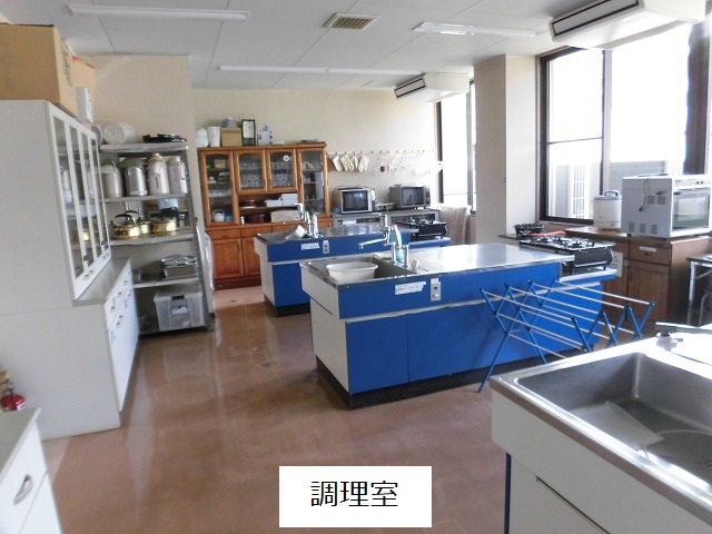 茶色い床と大きな窓が特徴の調理室で、中央には青と白の大きな調理台が二つ並んでおり、部屋の左奥には、食器棚と調理器具が収納された棚が並び、右奥の窓際には調理家電が置かれた調理室の写真