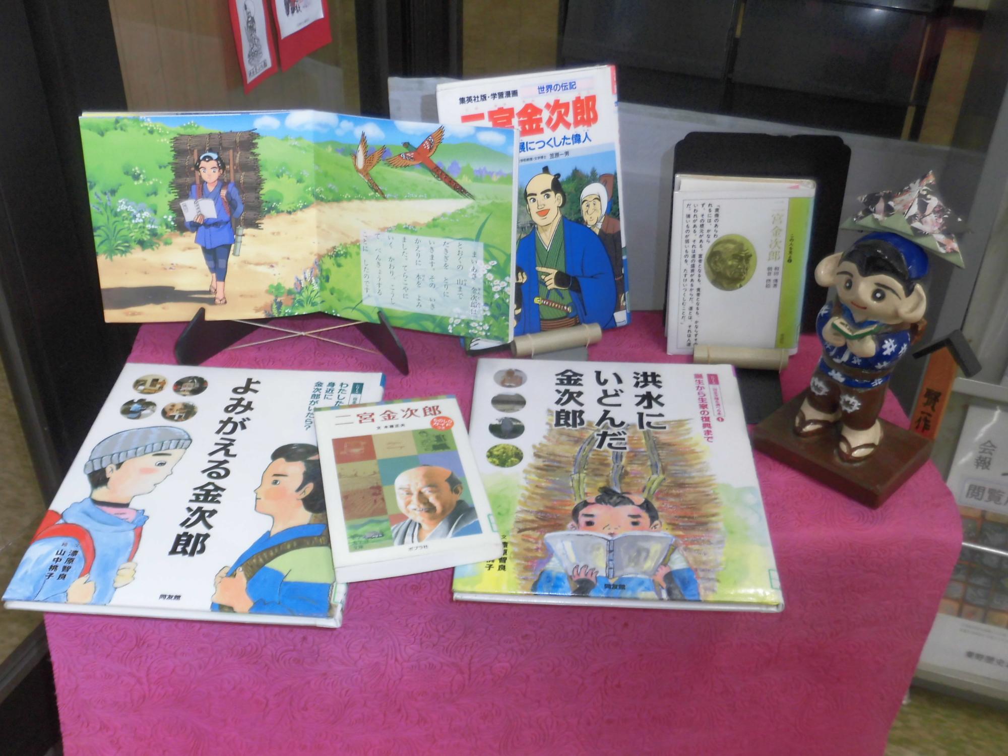 ピンクの布がかけられた台の上に、薪を背負って本を読む二宮金次郎のイラストが描かれた本や、彼の生涯を紹介する児童書、関連する人形が展示されている様子の写真