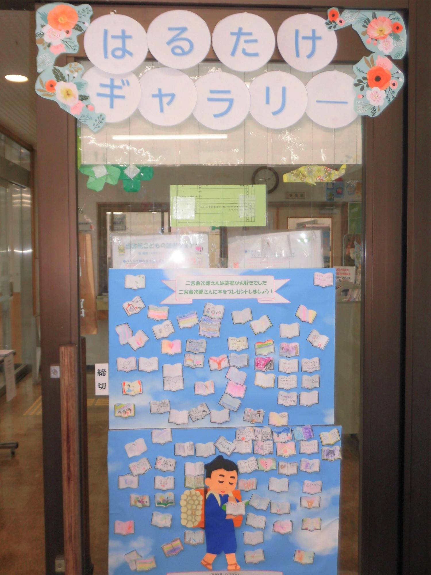 薪を背負って本を読む二宮金次郎のイラストが描かれた掲示板に、読書感想を書いた小さな本の形をした紙がたくさん貼られ、その上に1文字ずつ「はるたけギャラリー」と書かれた丸い紙が入のドアに貼られた写真