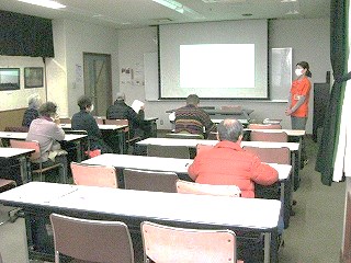 数名の受講者が机と椅子に座り、前方でプロジェクターに映し出された画面を指差しながら熱心に説明する女性講師の話を聞いている様子を捉えた写真