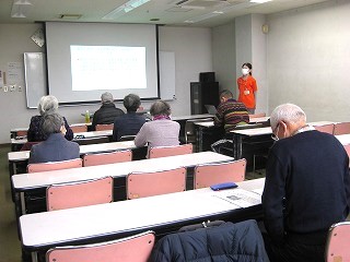 複数の受講者が机と椅子に座って、前方でプロジェクターに映し出された画面を見ながら女性講師の話を静かに聞いている様子を撮影した写真