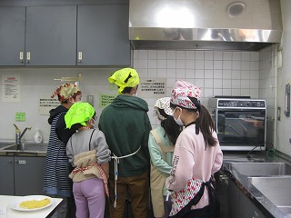 複数の人がエプロンと頭巾を着用し、厨房で調理台を囲んで何かの作業に取り組んでいる様子を後ろから撮影した写真