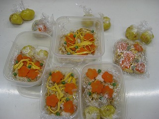 ラップで包まれた数個のサツマイモとリンゴの茶巾の横に、薄焼き卵と人参の飾り切りが乗ったちらし寿司の料理が透明なプラスチック容器に盛り付けられている様子を捉えた写真