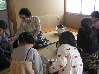 和室の畳の上で、数人の子供たちが座って見守る中、一人の女性が中央に置かれた道具を使ってお茶を立てている様子を捉えた写真