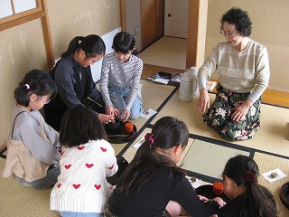 和室の畳の上で、数人の子供たちが座って見守る中、一人の女性が中央に置かれた茶道具を使ってお茶を立てている様子を捉えた写真