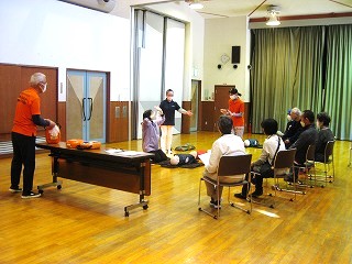 広い講堂で、数名の受講者が椅子に座り、前方で講師が心肺蘇生法のマネキンを使って実演指導を行っている様子を撮影した写真