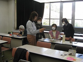 教室で数人の生徒が各自の机に向かう中、2人の女性が立ち、マスクを着用した別の2人の女性にスマホのような画面を見せて説明している様子を捉えた写真