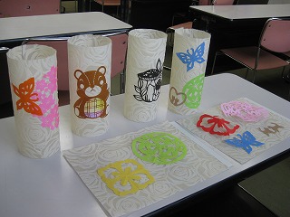 動物や蝶などの模様が切り抜かれた、薄い紙で作られたランタンと、それと同じデザインの切り絵作品が机の上に展示されている様子を撮影した写真