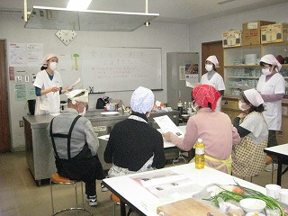 複数の参加者が机と椅子に座って講師の説明を聞き、別の参加者たちは講師と一緒に立っている様子を捉えた写真