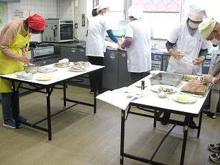 数人の参加者がそれぞれ台に分かれて立ち、手作り料理の盛り付けや準備作業を協力しながら行っている様子を捉えた写真