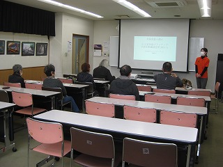 椅子に座った数名の受講者が、前方でプロジェクターに映し出された画面を指しながら説明する講師の話を静かに聞いている様子を撮影した写真