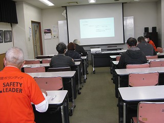 講習会で数名の受講者が机と椅子に座り、前方でプロジェクターに映し出された画面を見ながら真剣に講師の話を聞いている様子を捉えた写真