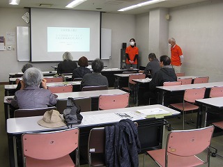 会議室のような部屋で、数名の受講者が机と椅子に座り、前方でプロジェクターに映された画面を指差しながら説明する女性講師の話を真剣に聞いている様子を撮影した写真