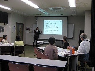 部屋の前方でスーツを着た講師がプロジェクターの画面を指しながら熱心に説明し、複数の受講者が机と椅子に座ってその講義を聞いている様子を捉えた写真