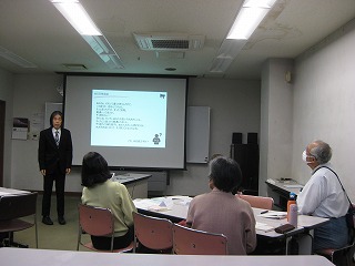 狭い部屋のプロジェクター画面の前でスーツを着た講師が講義を行い、数名の受講者が机と椅子に座って聴講している様子を写した写真